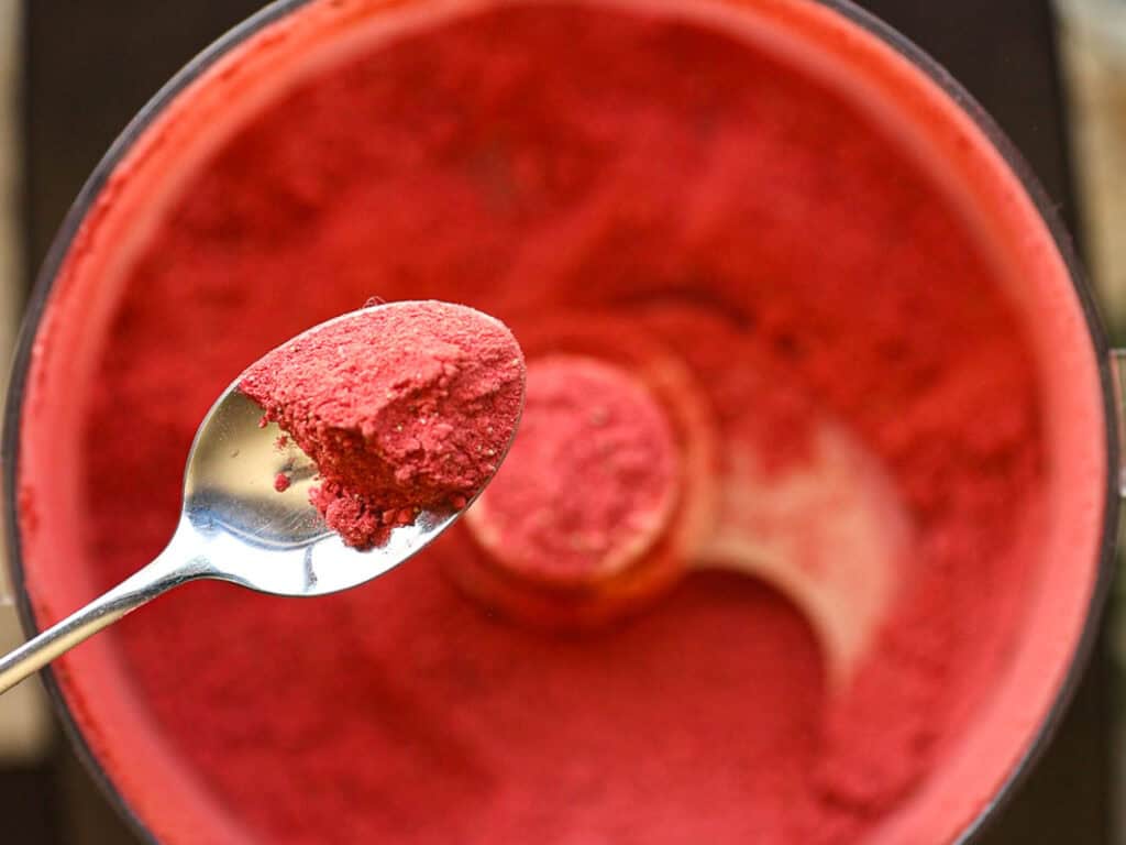 A close-up of a spoon holding bright red powder, hovering above a food processor filled with more of the same substance. The processors metal blade is visible amidst the vibrant powder.